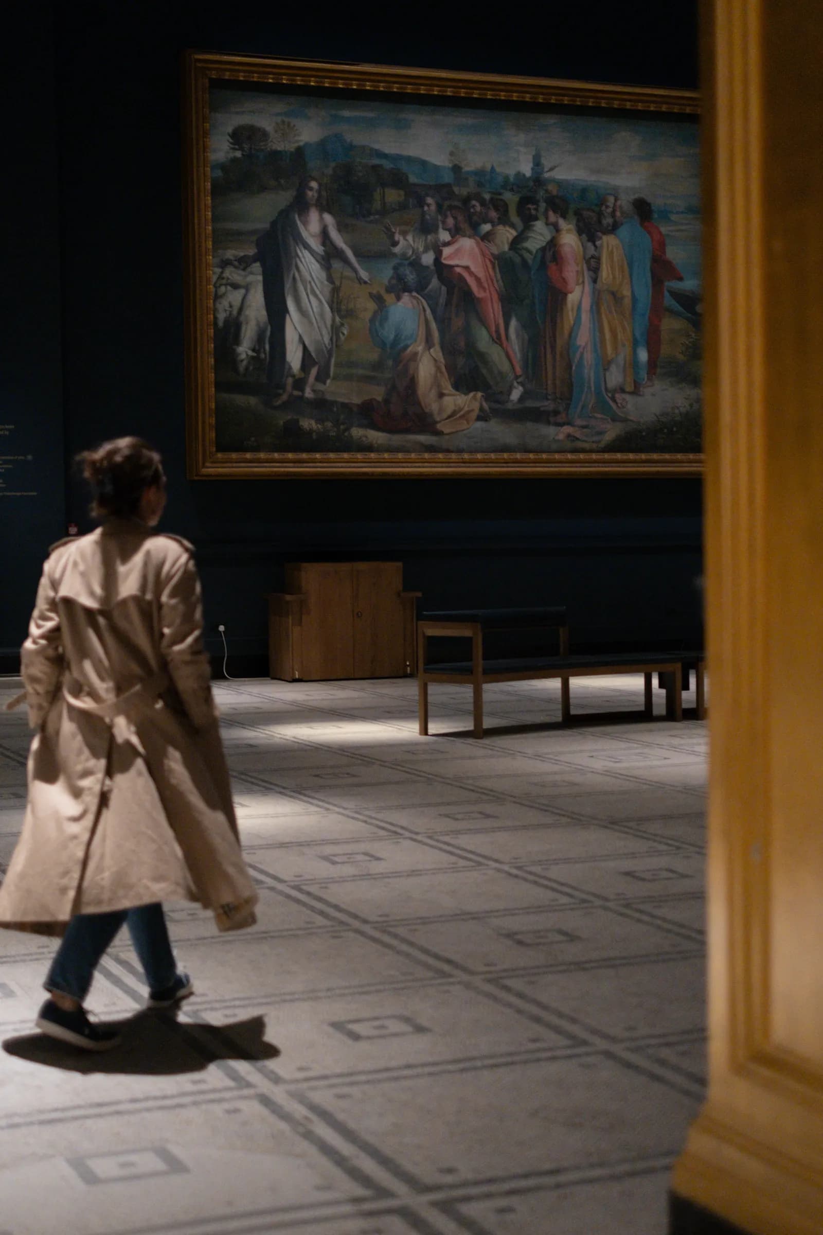 Woman in a trench walking across a mosaic floor beneath a vast old-master painting
